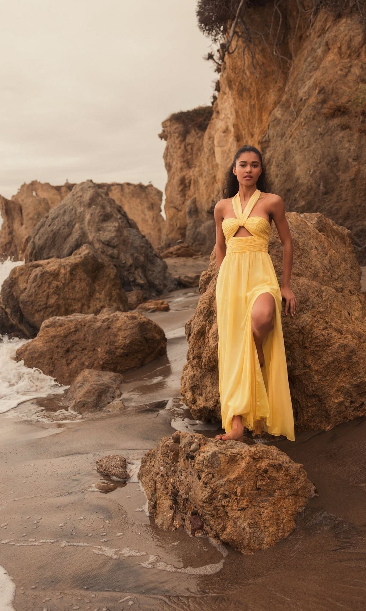 A person in the Long Formal Dress 55898 by Mac Duggal, featuring a yellow ruched halter neck design, stands on rocks by the ocean with cliffs, waves, and a cloudy sky in the background.